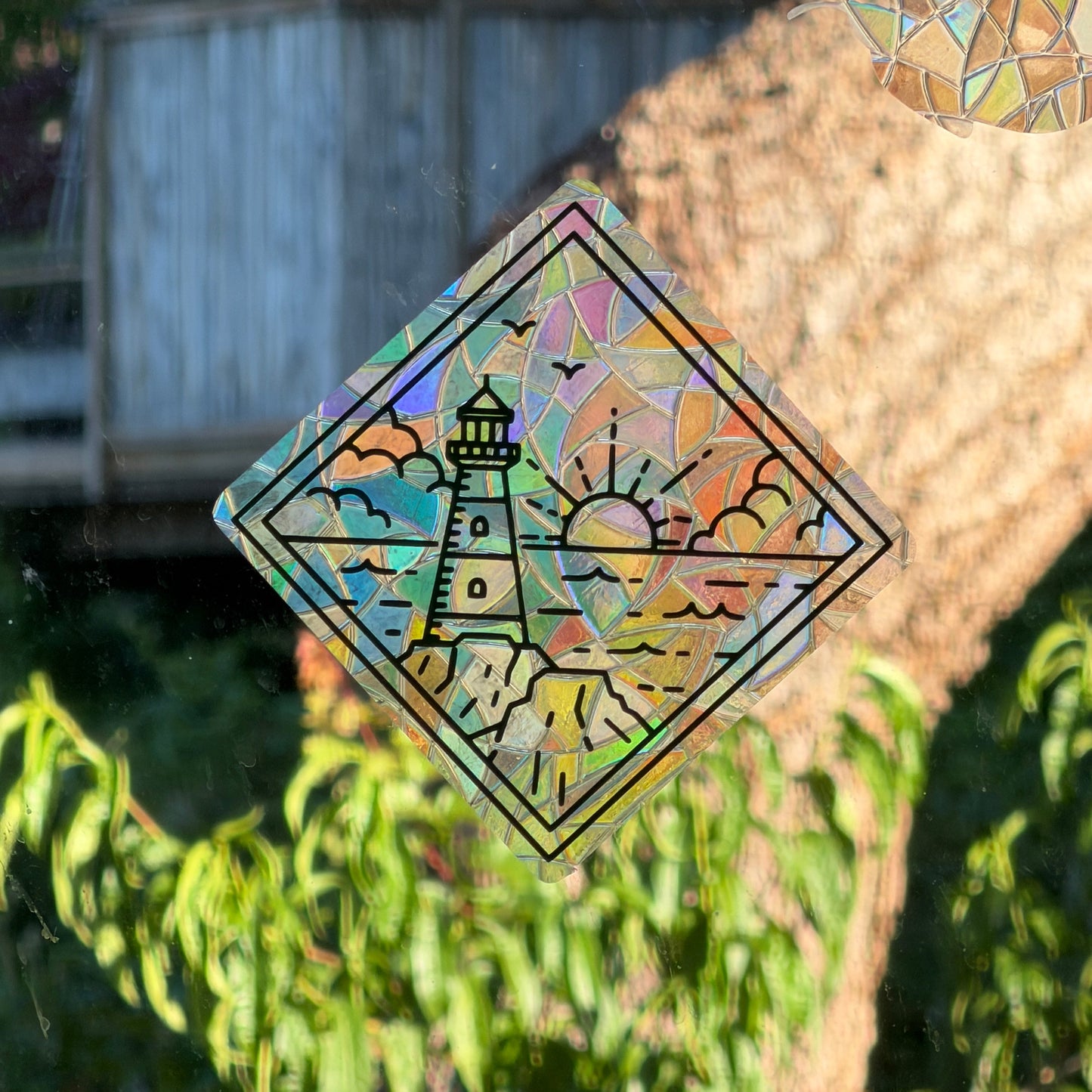 A diamond-shaped holographic sticker with a lighthouse and sun design, shown applied to a glass surface with a blurred outdoor background of greenery and a wooden structure.